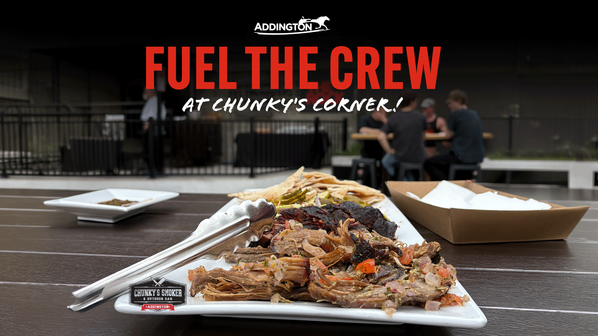 A large platter of shredded and charred meat with tongs, flatbread, and napkins sits on a wooden table in an outdoor dining area under a red "FUEL THE CREW" text overlay.