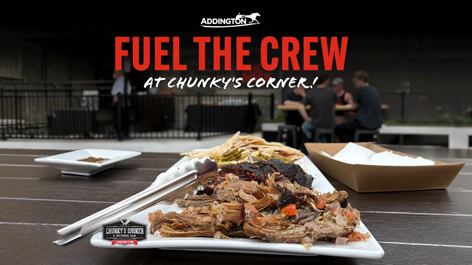 A large platter of shredded and charred meat with tongs, flatbread, and napkins sits on a wooden table in an outdoor dining area under a red "FUEL THE CREW" text overlay.