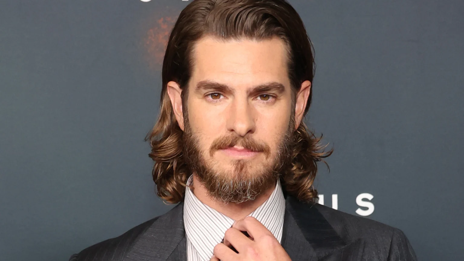 Andrew Garfield, with long brown hair and a full beard, looks forward while wearing a dark pinstripe suit and striped shirt, adjusting his collar.