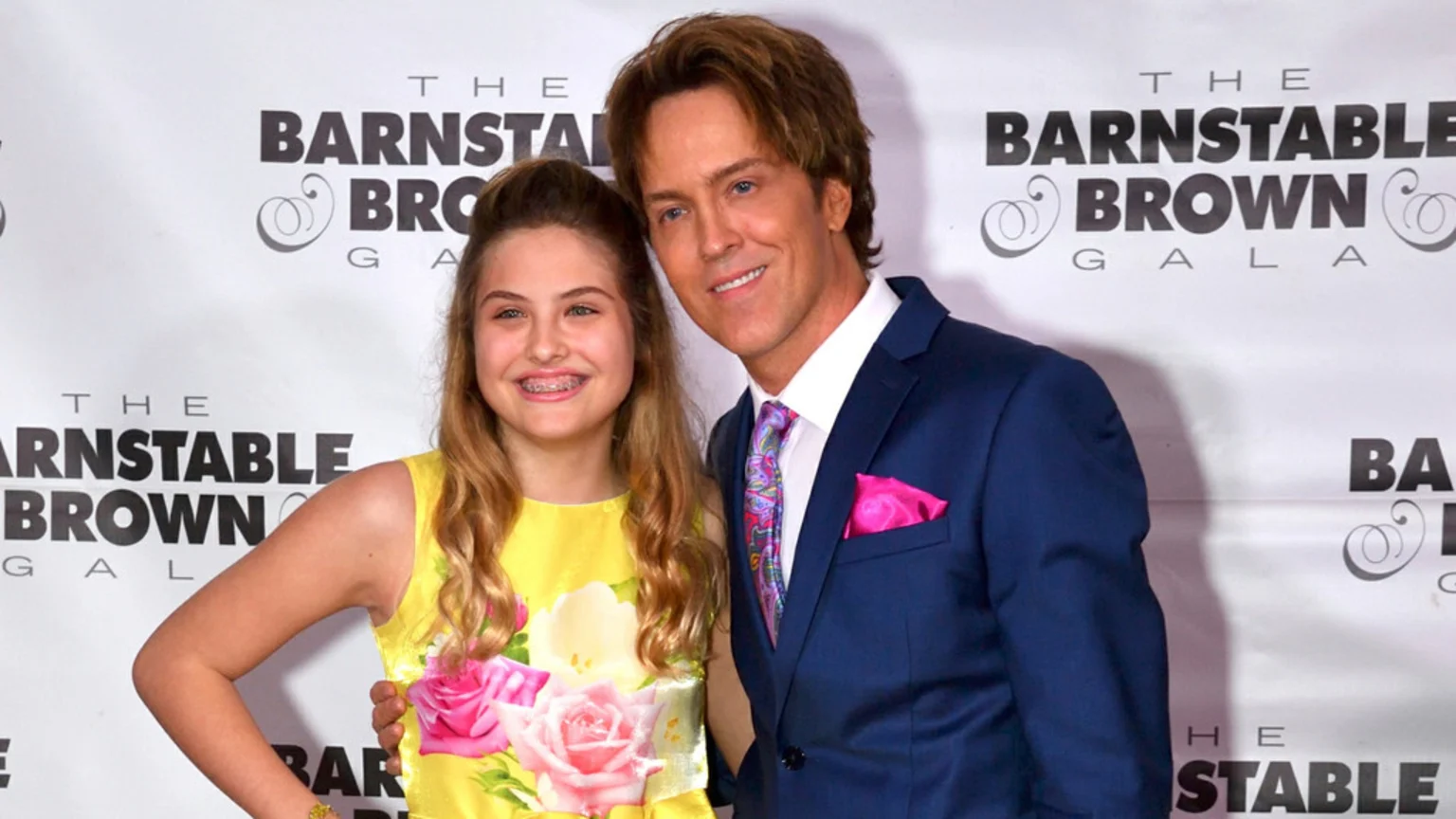 A man and a young girl smile at the camera, standing in front of a white event banner. The man wears a blue suit and colorful tie, while the girl wears a yellow floral dress and has braces.