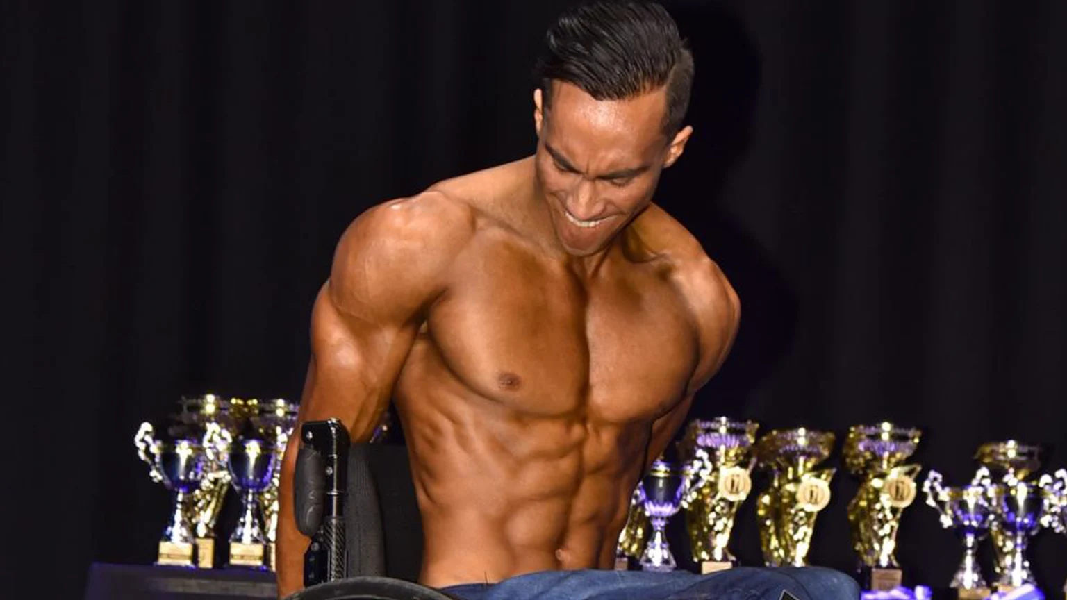 A smiling, muscular, dark-tanned man in a wheelchair poses shirtless on stage, flexing his torso with numerous gold and silver trophies displayed behind him.
