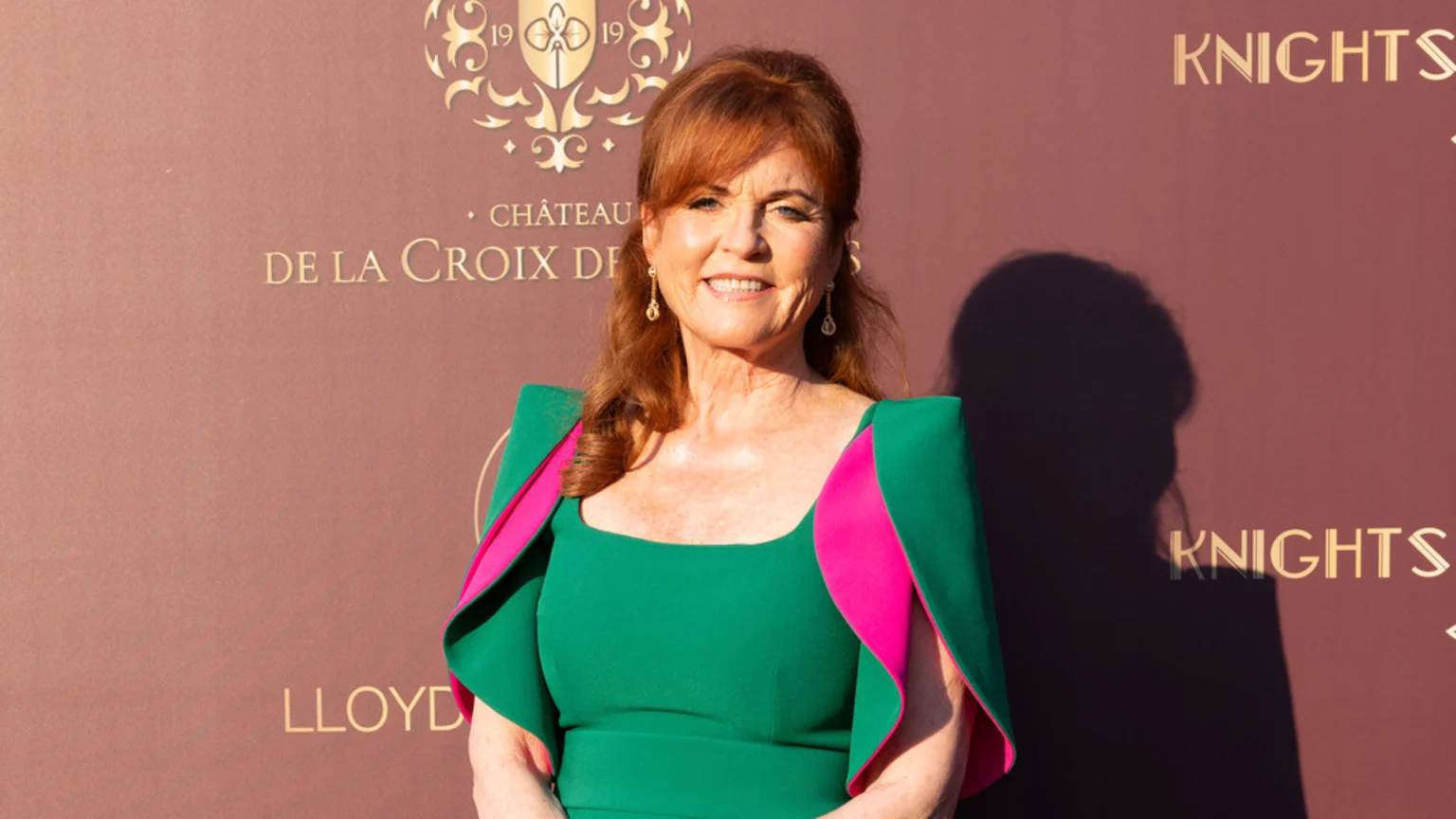 Sarah Ferguson smiles, wearing a green dress with pink-lined shoulders, standing in front of a reddish-brown wall with gold text.