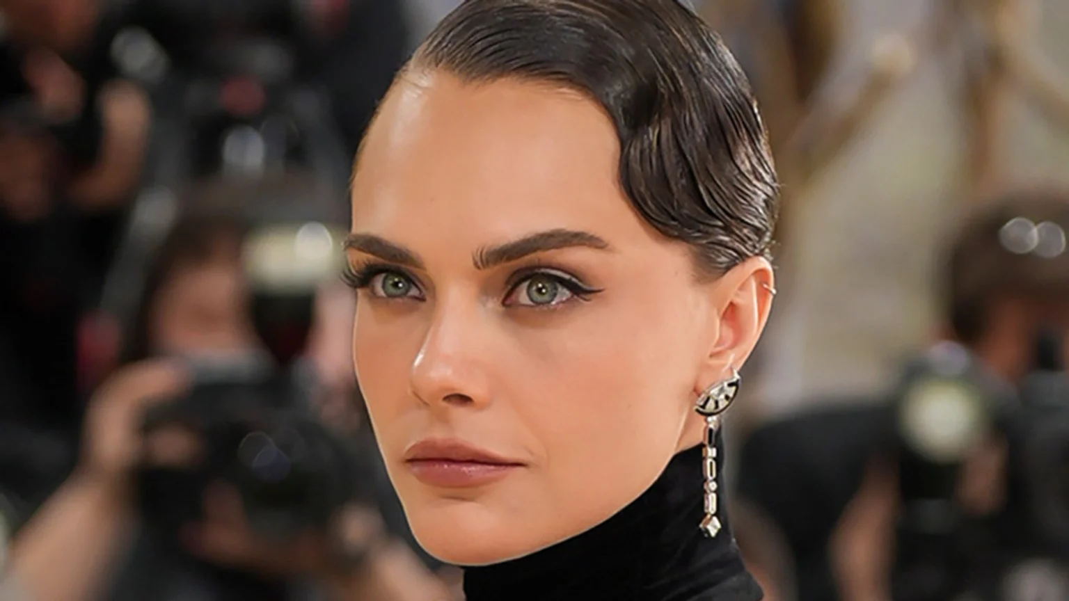 Close-up of Cara Delevingne with slicked-back dark hair styled in finger waves, light green eyes with dark eyeliner, and a decorative long earring, wearing a black turtleneck.