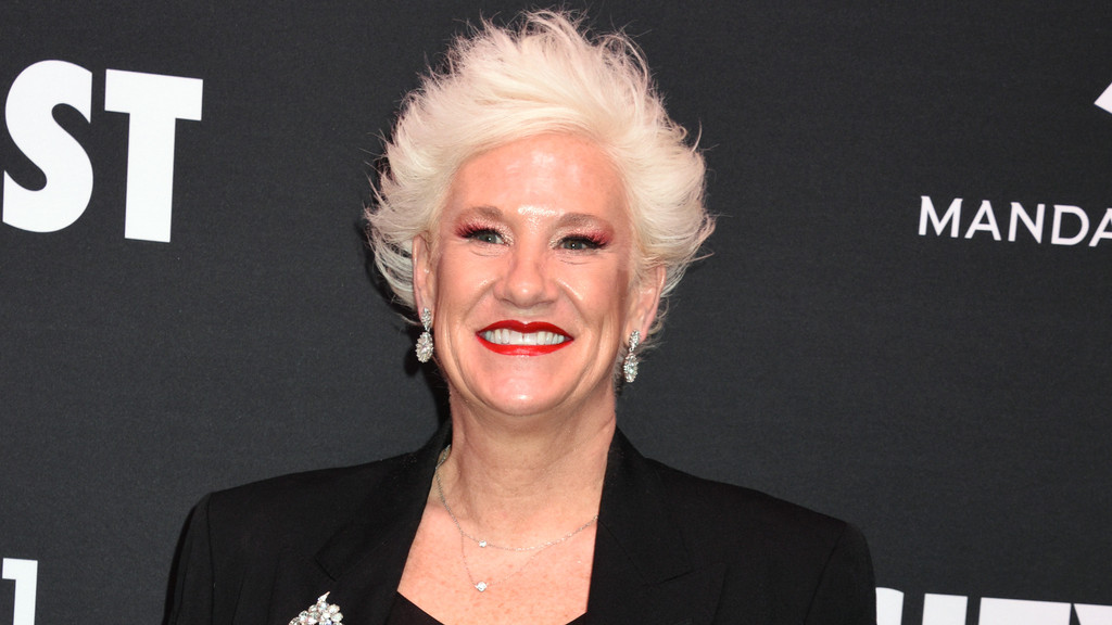 A close-up portrait of an unknown adult with short, spiky white hair, red lipstick, and red eyeshadow, smiling broadly against a dark event backdrop with white text. They wear a black blazer, silver necklace with clear stone pendants, dangling ornate earrings, and a sparkling brooch.