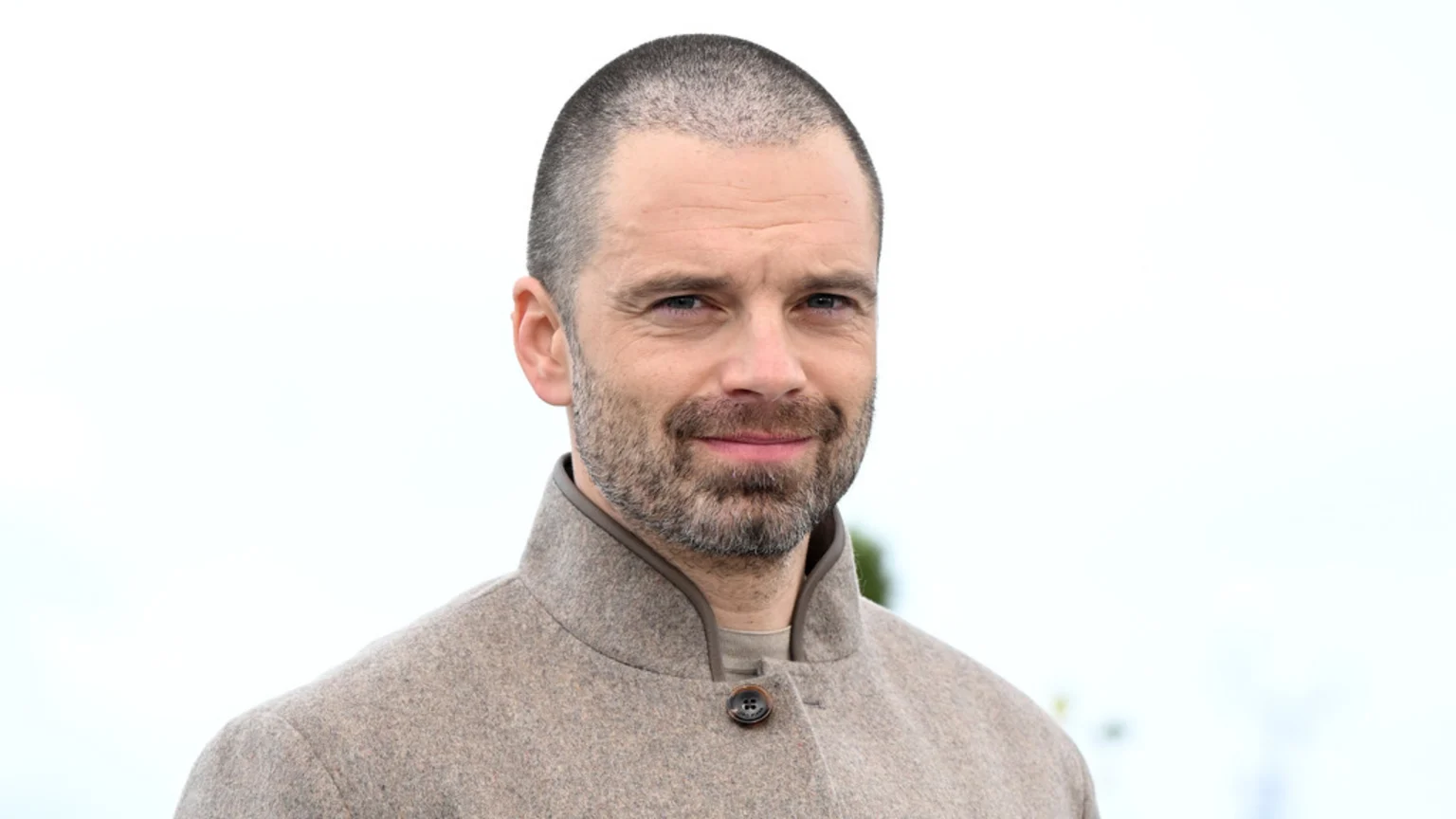 Sebastian Stan with a shaved head and beard, wearing a beige jacket, looking forward against a light background.