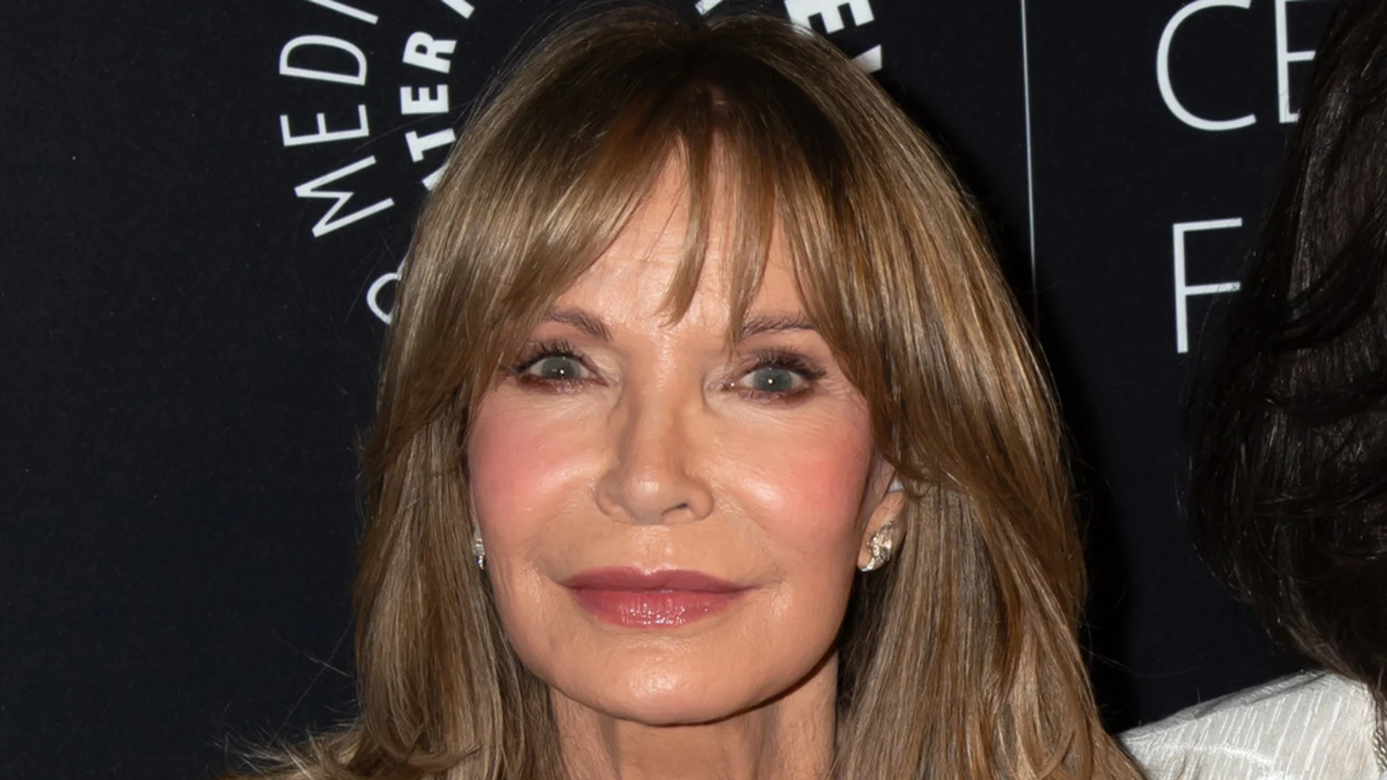Close-up of Jaclyn Smith with light brown hair and bangs, wearing makeup and earrings, looking towards the camera against a dark background with partial white text.