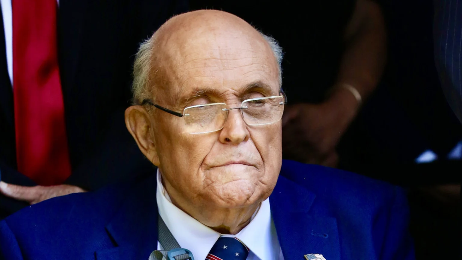 A close-up shows an older man with thinning white hair and glasses, wearing a blue suit and a patterned tie, with a serious expression.