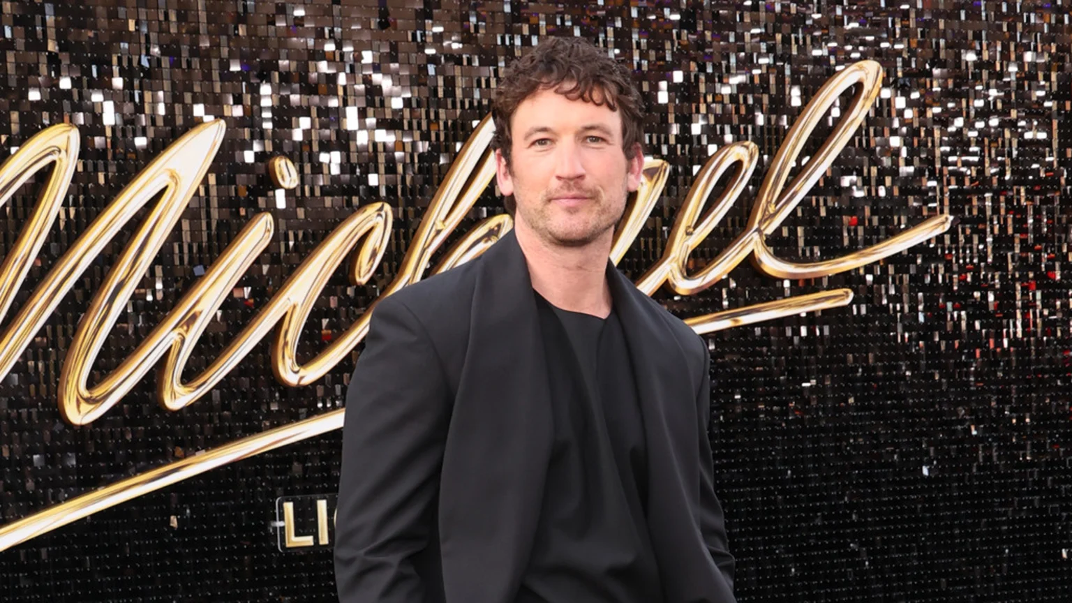 Miles Teller, with curly brown hair and a light beard, stands against a glittering black wall adorned with golden cursive letters spelling 'Michael', wearing a black blazer and t-shirt, looking at the camera.