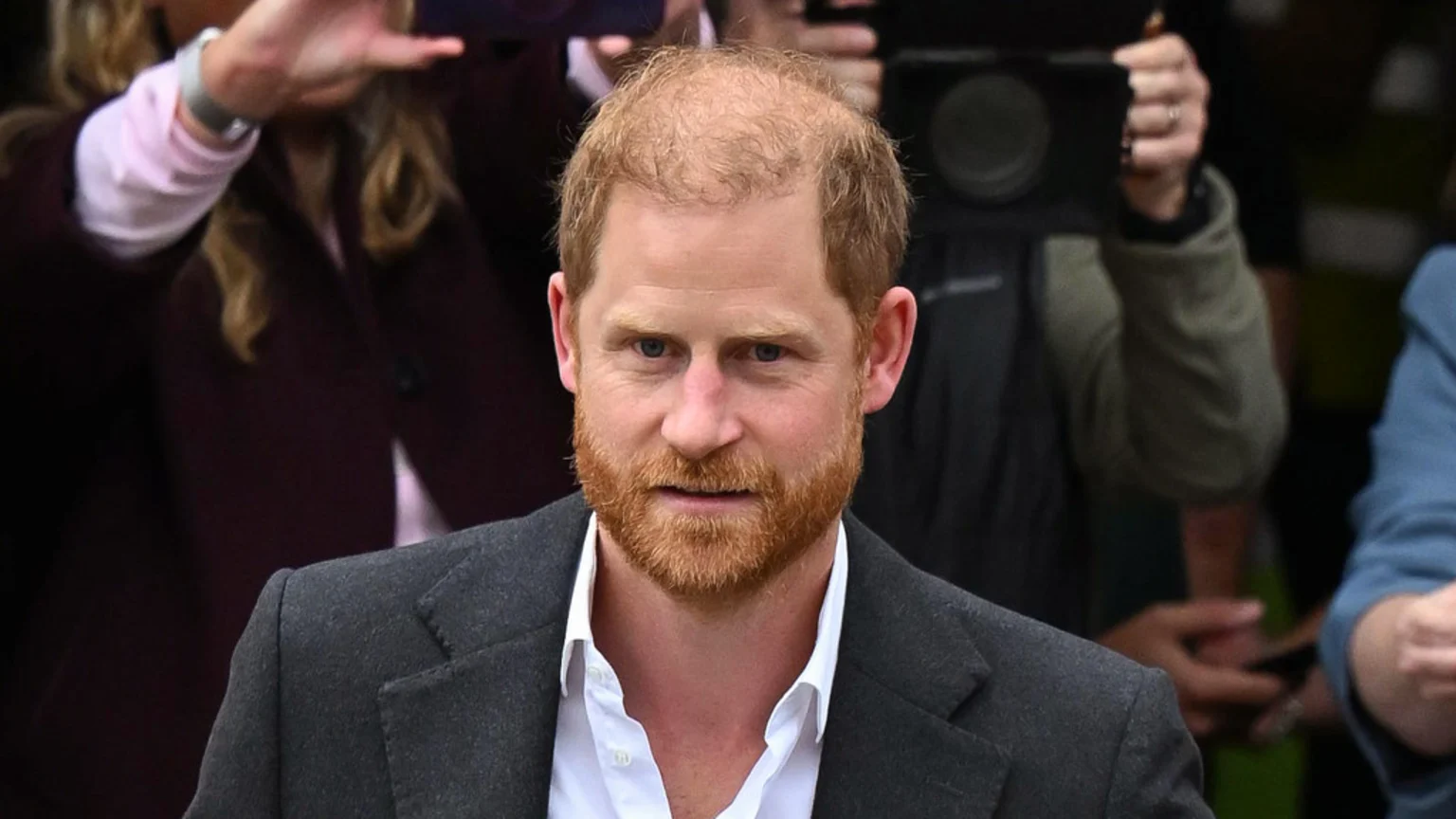 Prince Harry, with reddish hair and beard, looks forward with a serious expression, wearing a dark grey jacket and white shirt, with blurred figures and cameras in the background.