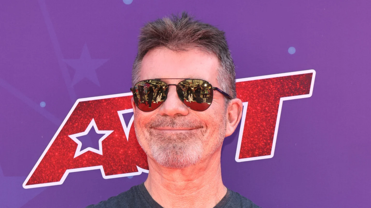 Simon Cowell, a gray-haired man with a beard, smiles in aviator sunglasses reflecting a crowd, against an America's Got Talent logo.