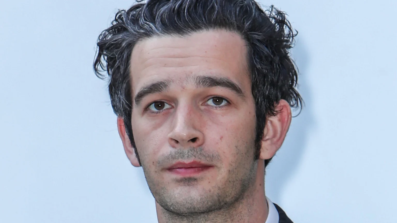 Close-up portrait of a man with messy dark hair with gray strands, brown eyes, and light stubble, looking directly at the viewer against a plain light background.