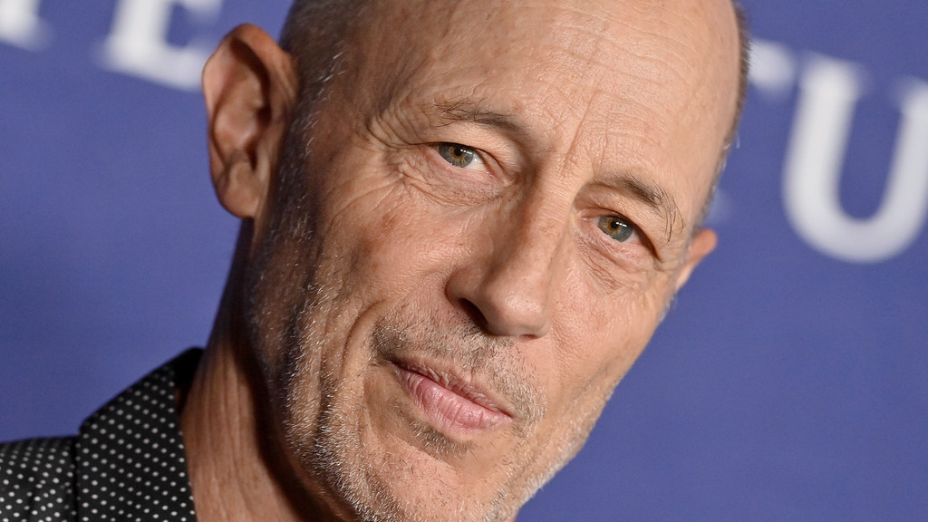 Close-up of a man with light skin, visible wrinkles, hazel eyes, a bald head, and facial stubble, wearing a black polka dot shirt against a blurry blue background with a white letter.