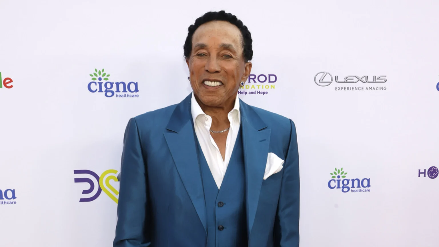 Smokey Robinson, smiling and wearing a blue suit and white shirt, stands in front of a white backdrop with various sponsor logos.