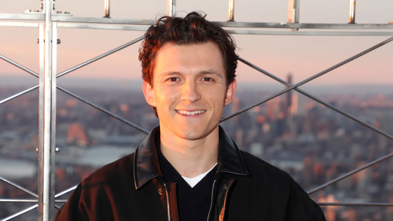 Actor Tom Holland smiles at the camera, wearing a dark jacket and sweater, against a blurred city skyline and metal railing bathed in warm, sunset light.
