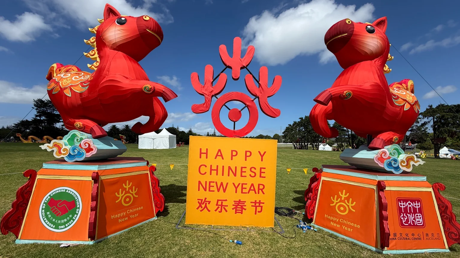 Two large red horse lanterns on decorated pedestals flank a yellow sign that reads "HAPPY CHINESE NEW YEAR 欢乐春节" at an outdoor event under a blue sky.