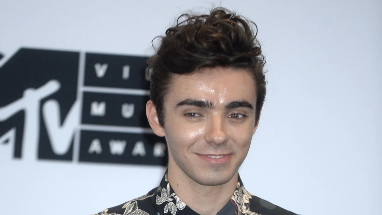 Nathan Sykes, a young man with curly dark hair and a patterned shirt, smiles in front of a blurred MTV Video Music Awards logo.