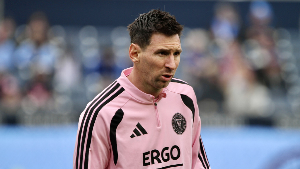 Lionel Messi in a pink Inter Miami training top with black stripes and logos, looking focused on a field with a blurred background.