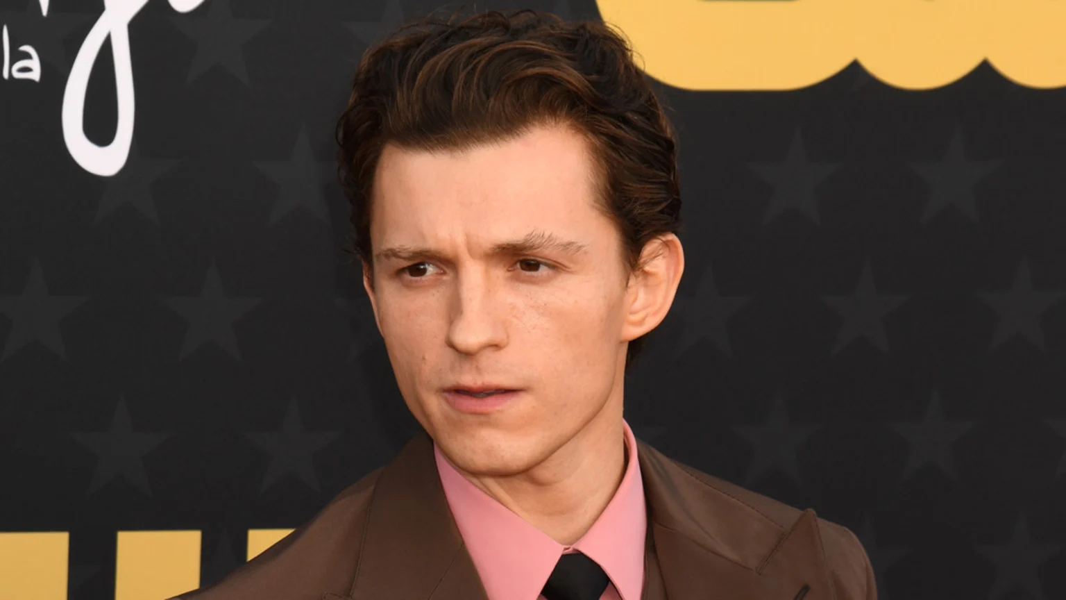 A close-up of actor Tom Holland, wearing a brown suit, pink shirt, and black tie, looking intently to his left against a dark, star-patterned background.