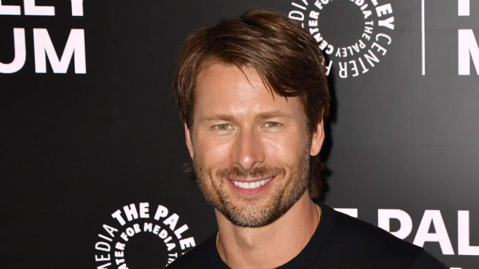 A smiling man with brown hair and a beard, wearing a dark shirt and gold chain, looks at the camera against a black backdrop with white Paley Center for Media logos.