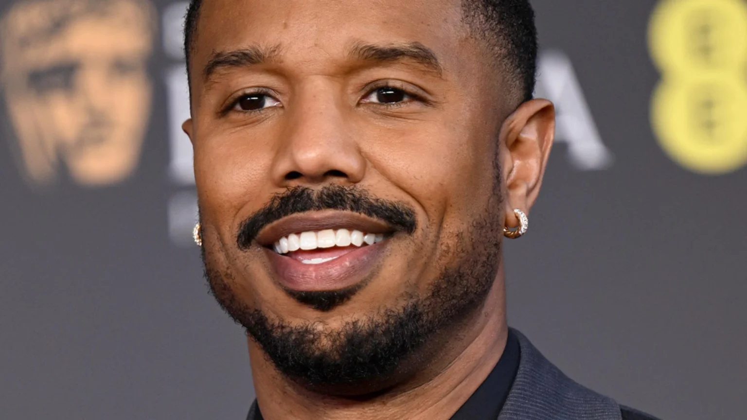 Close-up of Michael B. Jordan smiling widely, showing his teeth, with a beard, mustache, and diamond hoop earrings, against a blurred dark background.
