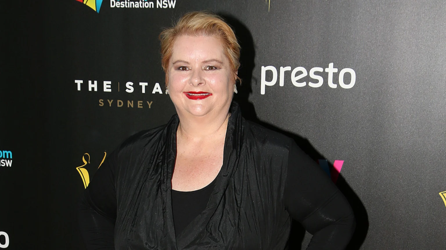 A smiling woman with short blonde hair and red lipstick wears a black top and vest, standing in front of a dark backdrop with event logos.