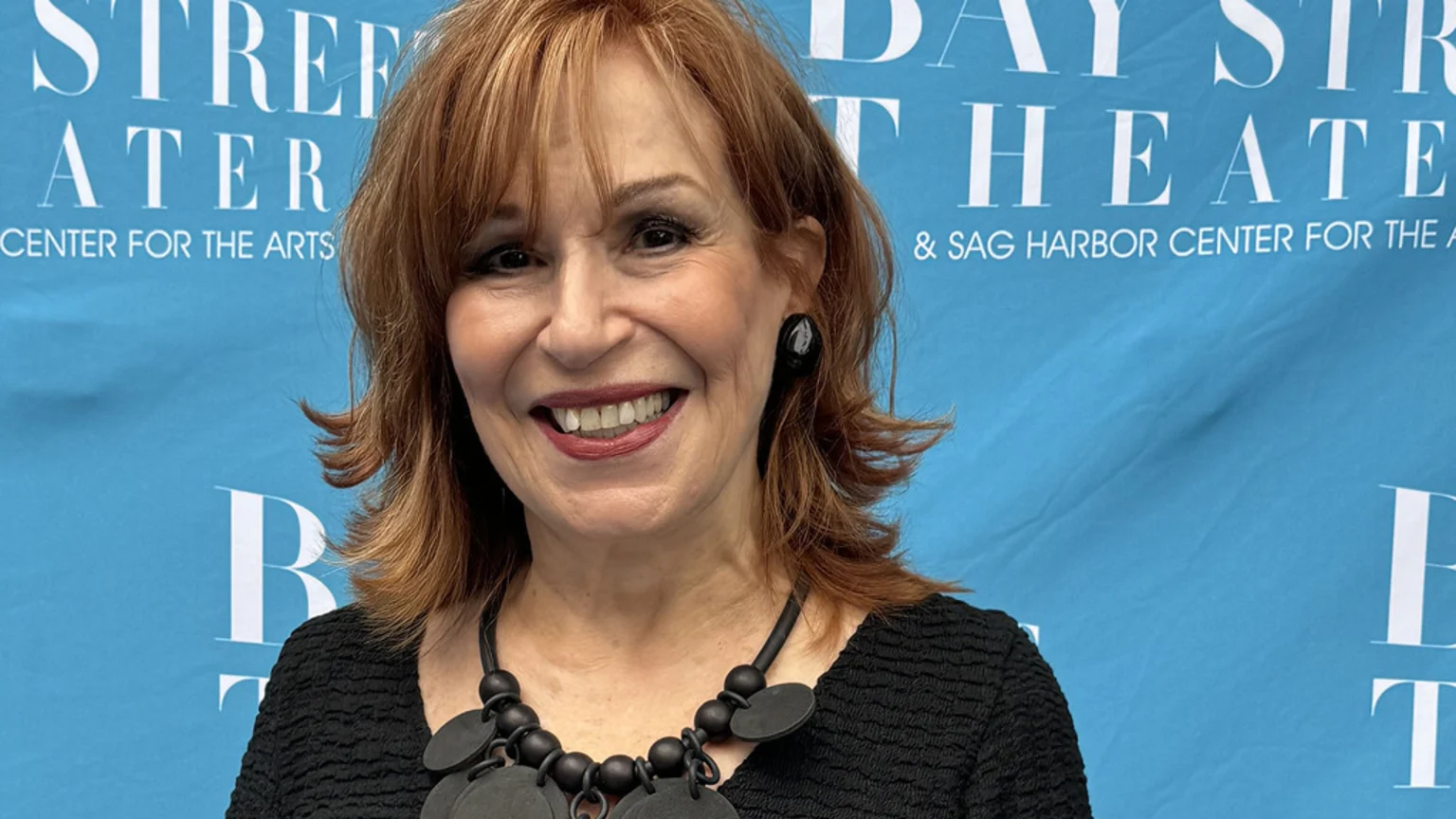 Joy Behar smiles broadly, wearing a black textured top and chunky black necklace, against a blue banner for the Bay Street Theater.
