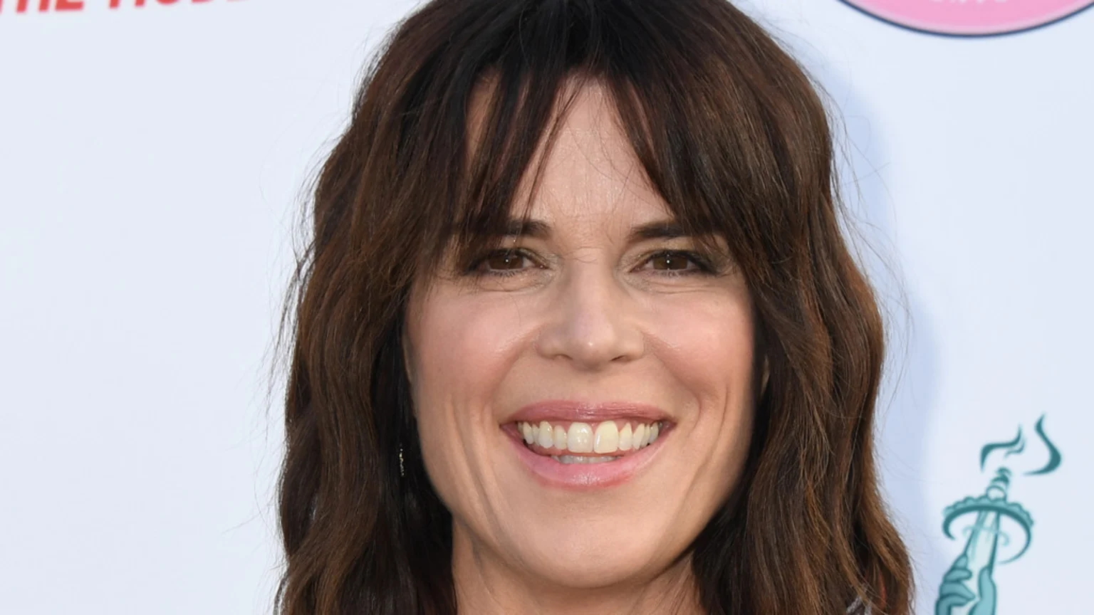 Close-up of Neve Campbell smiling and looking at the camera, with dark brown hair, bangs, and teeth visible.