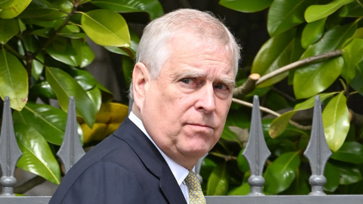 Prince Andrew, wearing a dark suit, white shirt, and yellow patterned tie, looks directly at the viewer with a serious expression, with green foliage and a grey fence in the background.