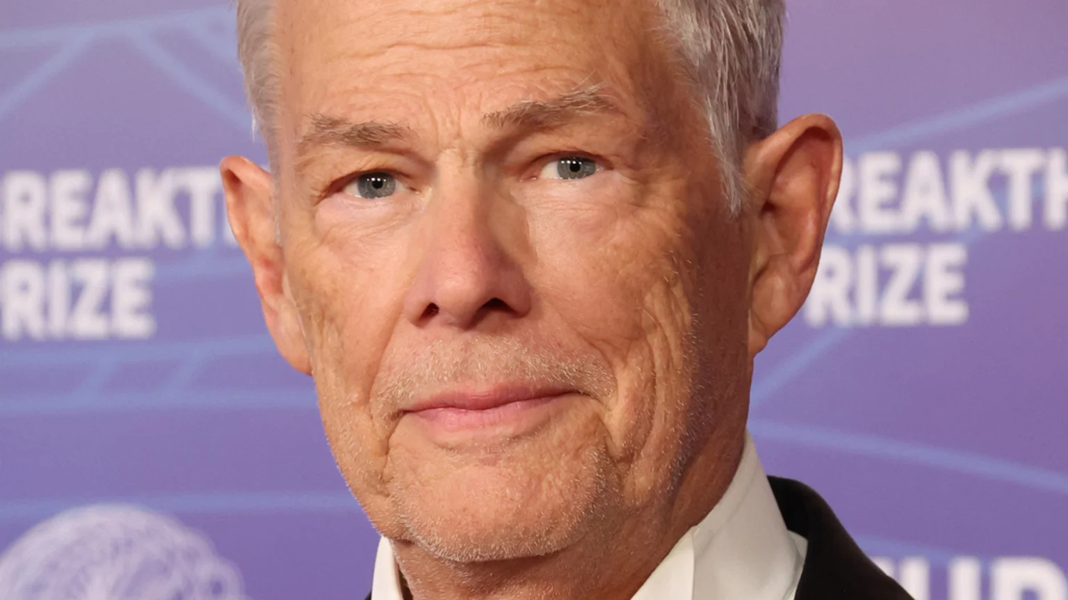 Close-up portrait of an older man with gray hair and blue eyes, wearing a white shirt and black suit, against a blurred purple background with white "BREAKTHROUGH PRIZE" text.