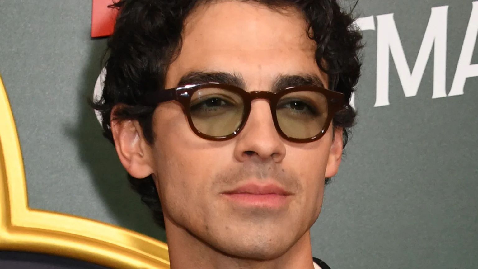 Close-up of Joe Jonas with dark curly hair and light stubble, wearing dark brown glasses with greenish-yellow tinted lenses, looking forward against a blurry greenish-gray background with white text.