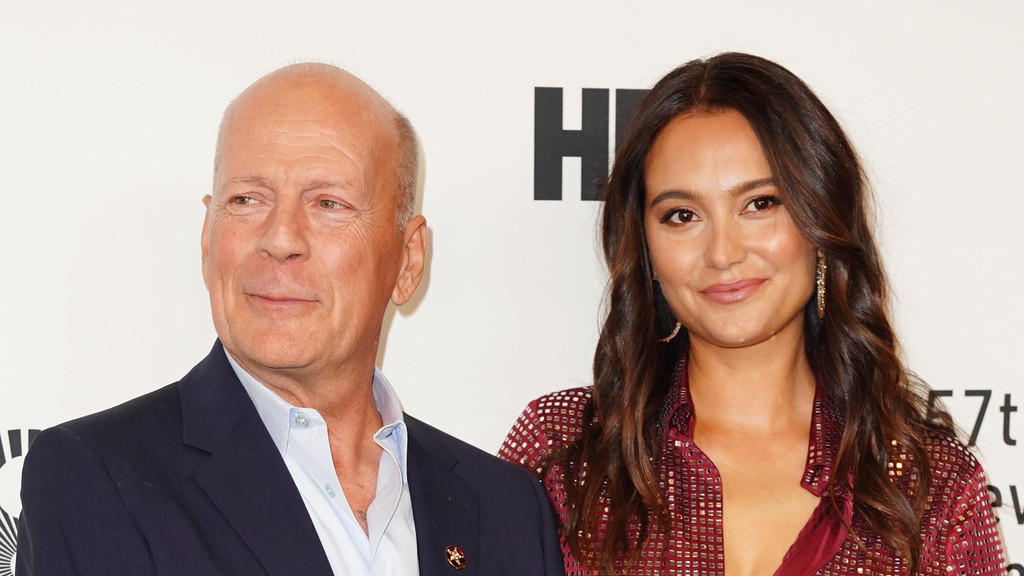 Bruce Willis, bald and in a dark suit, stands smiling next to a woman with long, dark, wavy hair and a patterned red top, who smiles at the viewer.