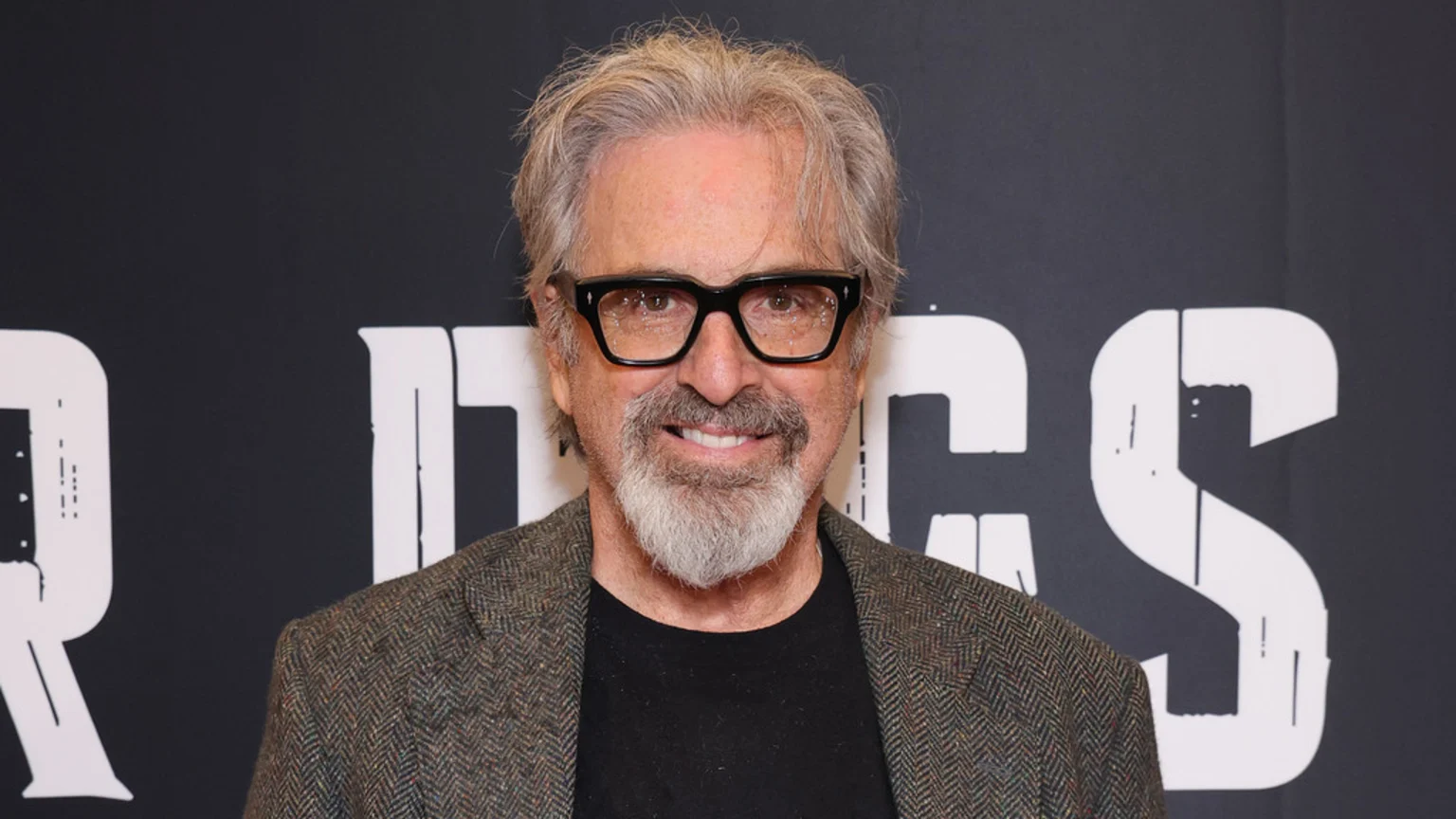Robert Carradine, a smiling man with grey hair, a beard, and black glasses, wears a herringbone jacket against a dark background with white letters.