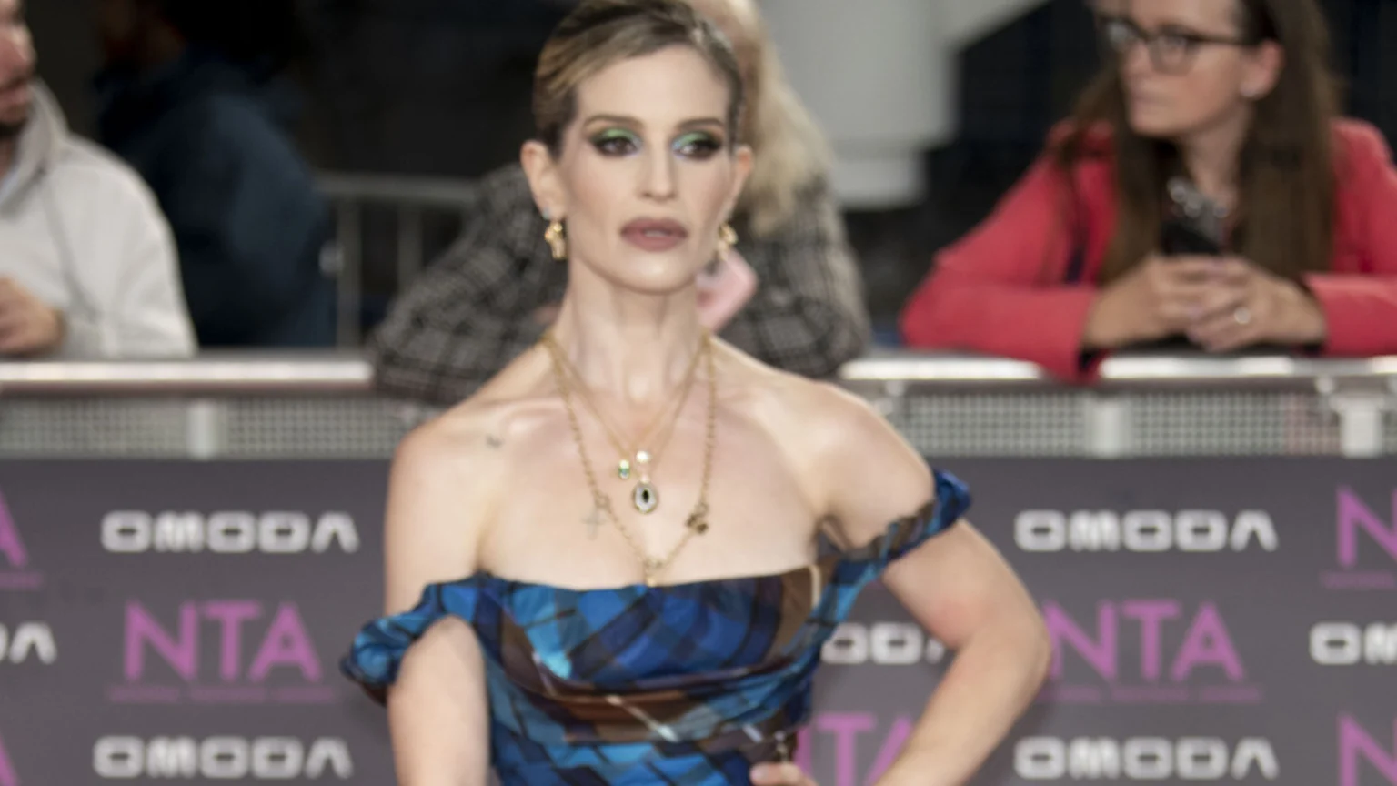 A woman with blonde hair, green and blue eye makeup, and gold necklaces wears an off-the-shoulder plaid dress in front of an "OMODA NTA" branded backdrop.
