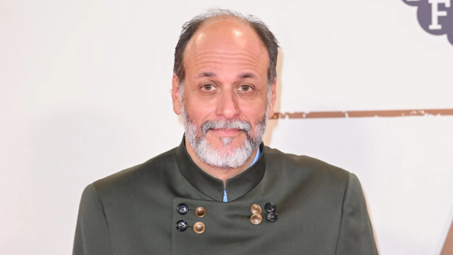 A man with a grey beard and dark hair, wearing a dark olive green jacket with a high collar and decorative buttons, looks directly at the camera.