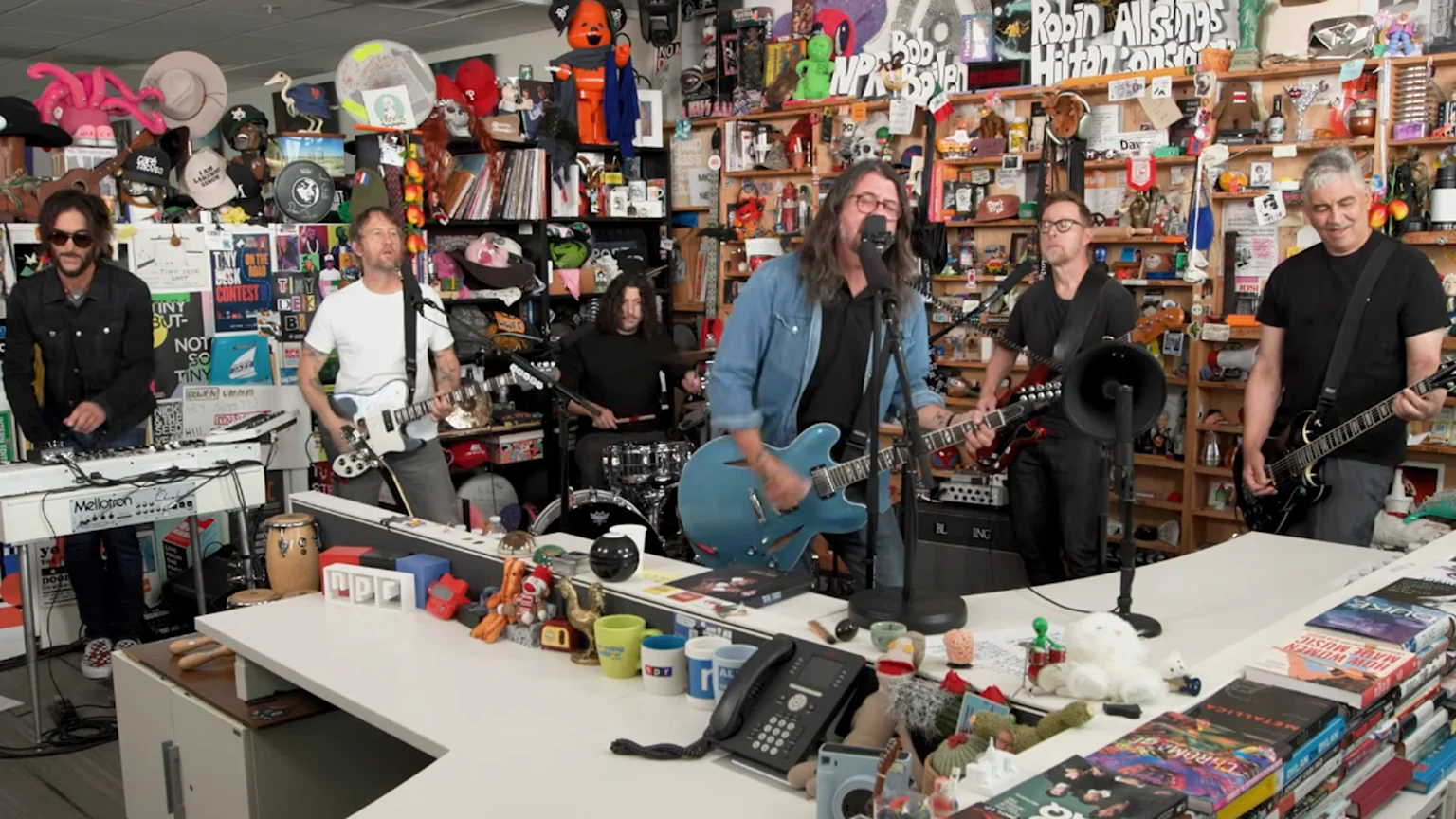 Six musicians perform in a cluttered office with a desk full of items, featuring a lead singer on a blue guitar, a drummer, a keyboardist, and three other guitarists.