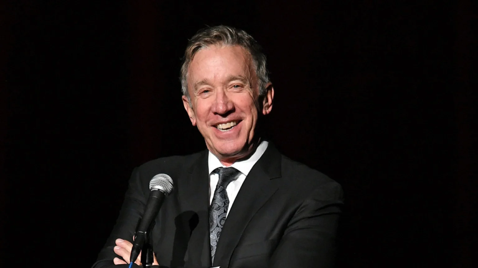 Tim Allen smiles broadly, looking to his left, while standing at a microphone with his hands clasped, wearing a dark suit and patterned tie against a black background.