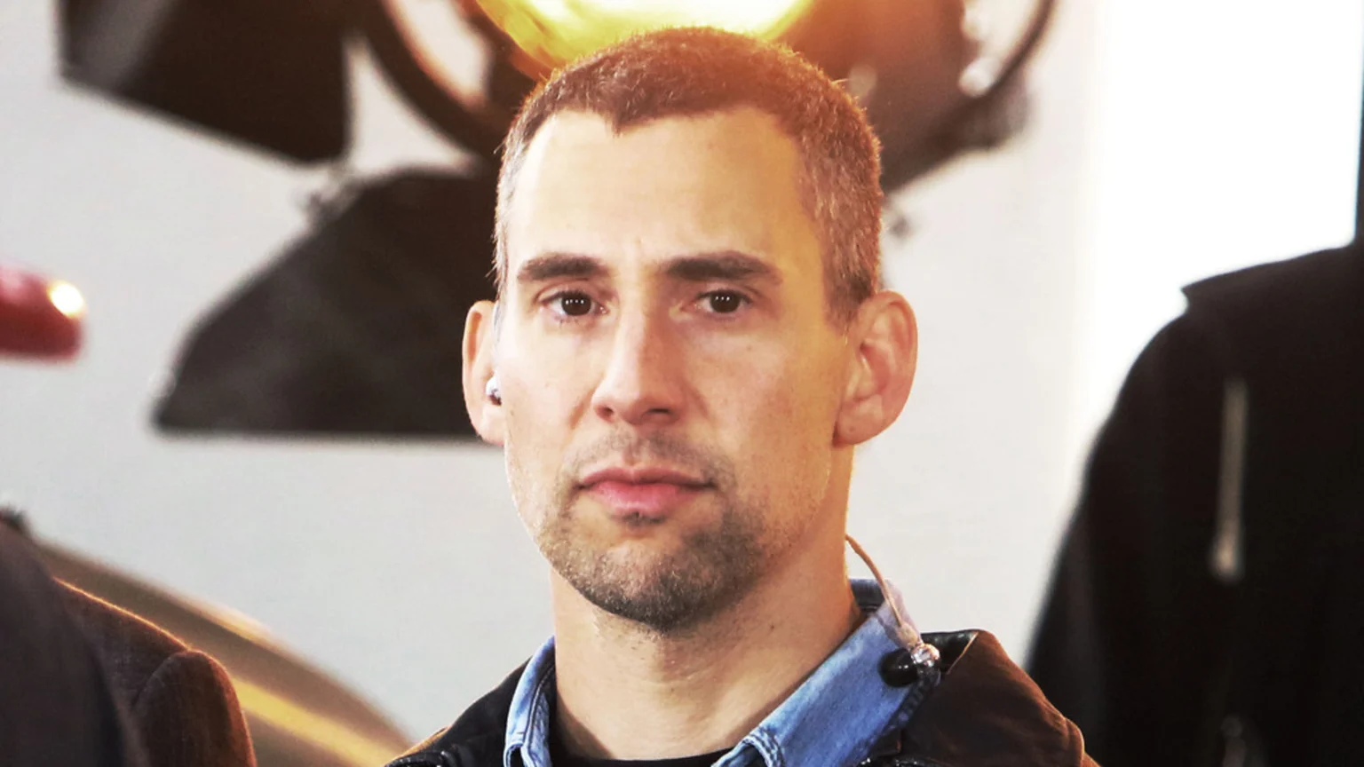 A man with short dark hair and stubble, lit by warm light, looks directly at the viewer, wearing a dark jacket over a denim shirt, with an earbud in his left ear.