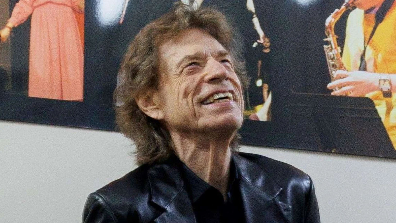 Mick Jagger smiles widely and looks upwards, wearing a black jacket, against a dark background with blurred figures including a person playing a saxophone.