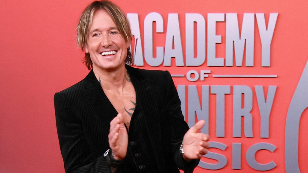 Keith Urban, a smiling man with blonde hair and visible tattoos, claps in front of a red background featuring the "Academy of Country Music" logo.