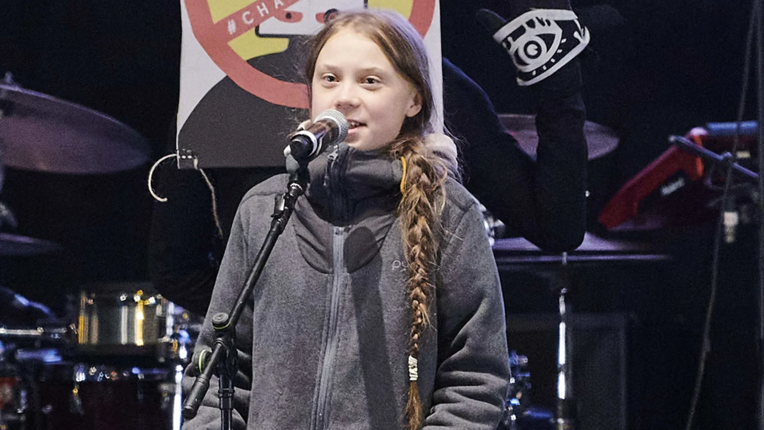 Greta Thunberg, a young woman with a long braid and grey jacket, speaks into a microphone on a stage with musical equipment in the background.