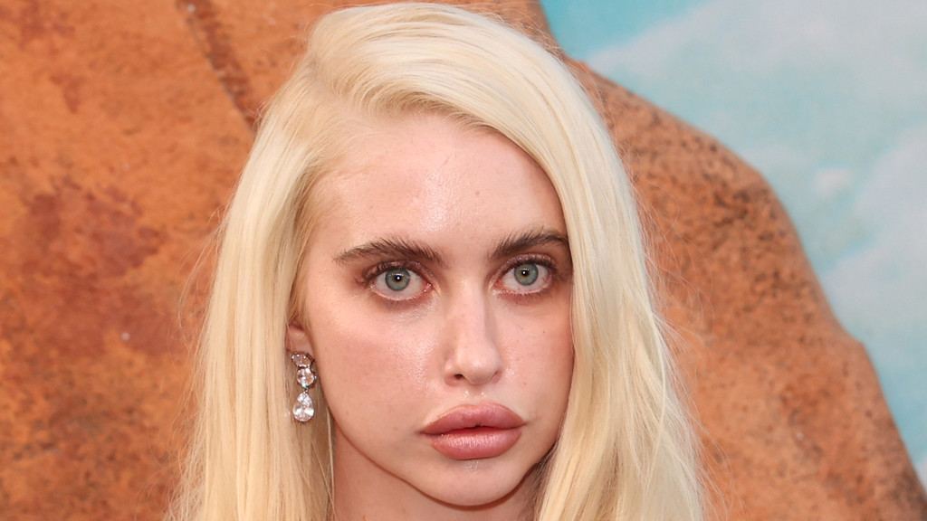 Close-up of actress Chloe Cherry with long blonde hair, light blue eyes, prominent eyebrows, and full lips, wearing a dangling clear gemstone earring.