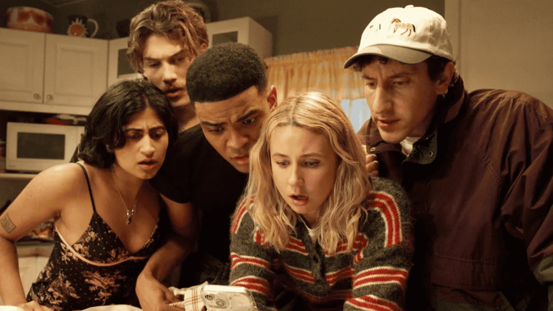 Five people with surprised and focused expressions gather closely around a blonde woman holding a smartphone, intently looking at its screen in a home setting.