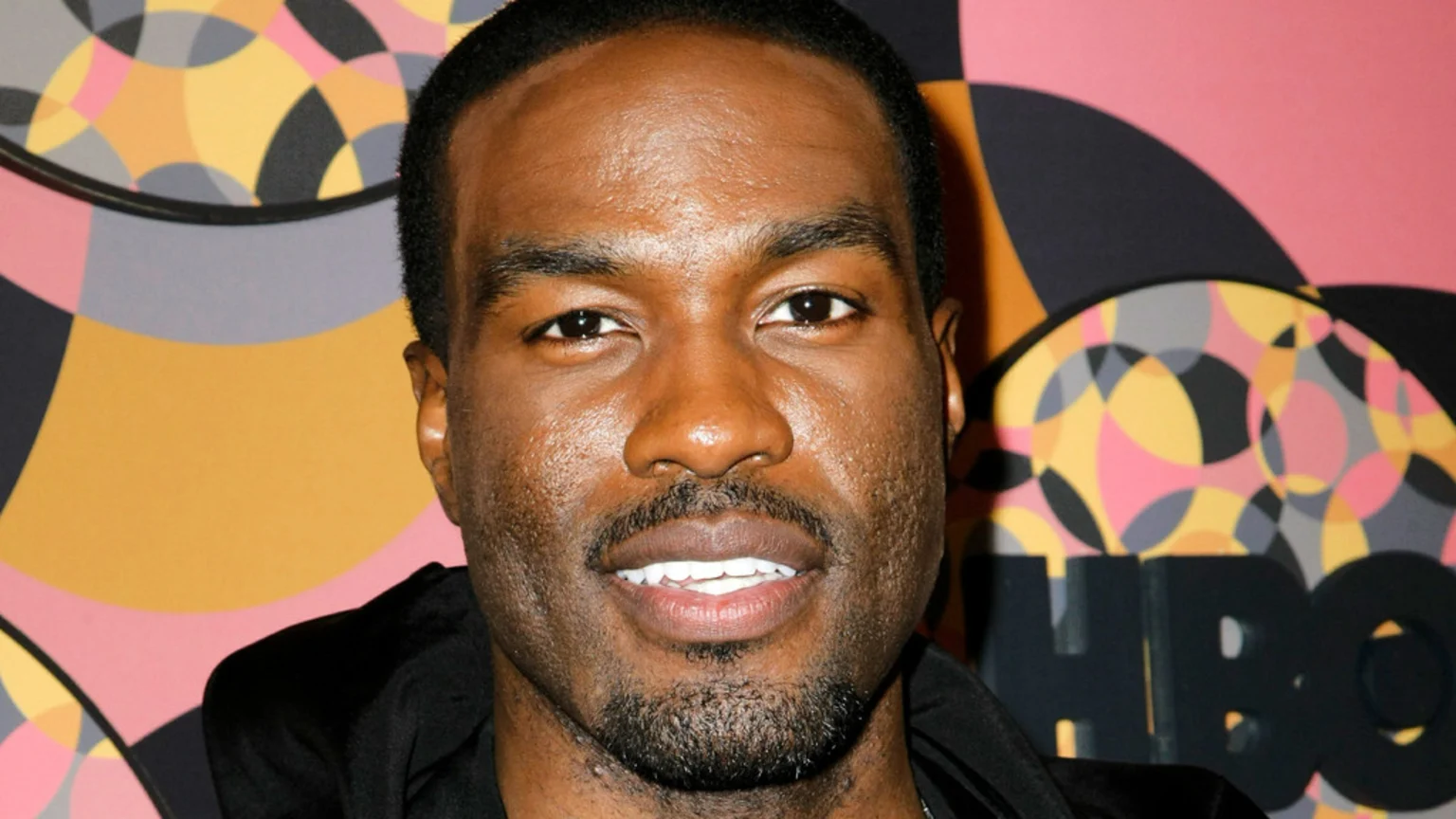 A close-up portrait of actor Yahya Abdul-Mateen II smiling slightly, with a colorful geometric background that includes the HBO logo.