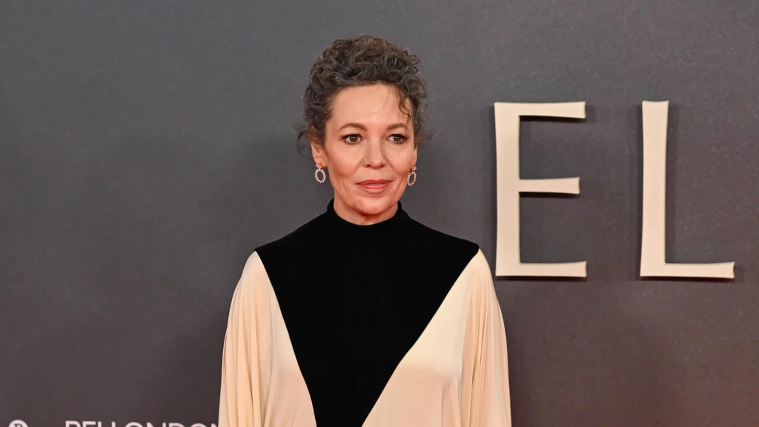 Olivia Colman, with curly hair pulled back, wears a black and cream color-block dress and drop earrings, standing against a dark grey background with letters "EL" visible.