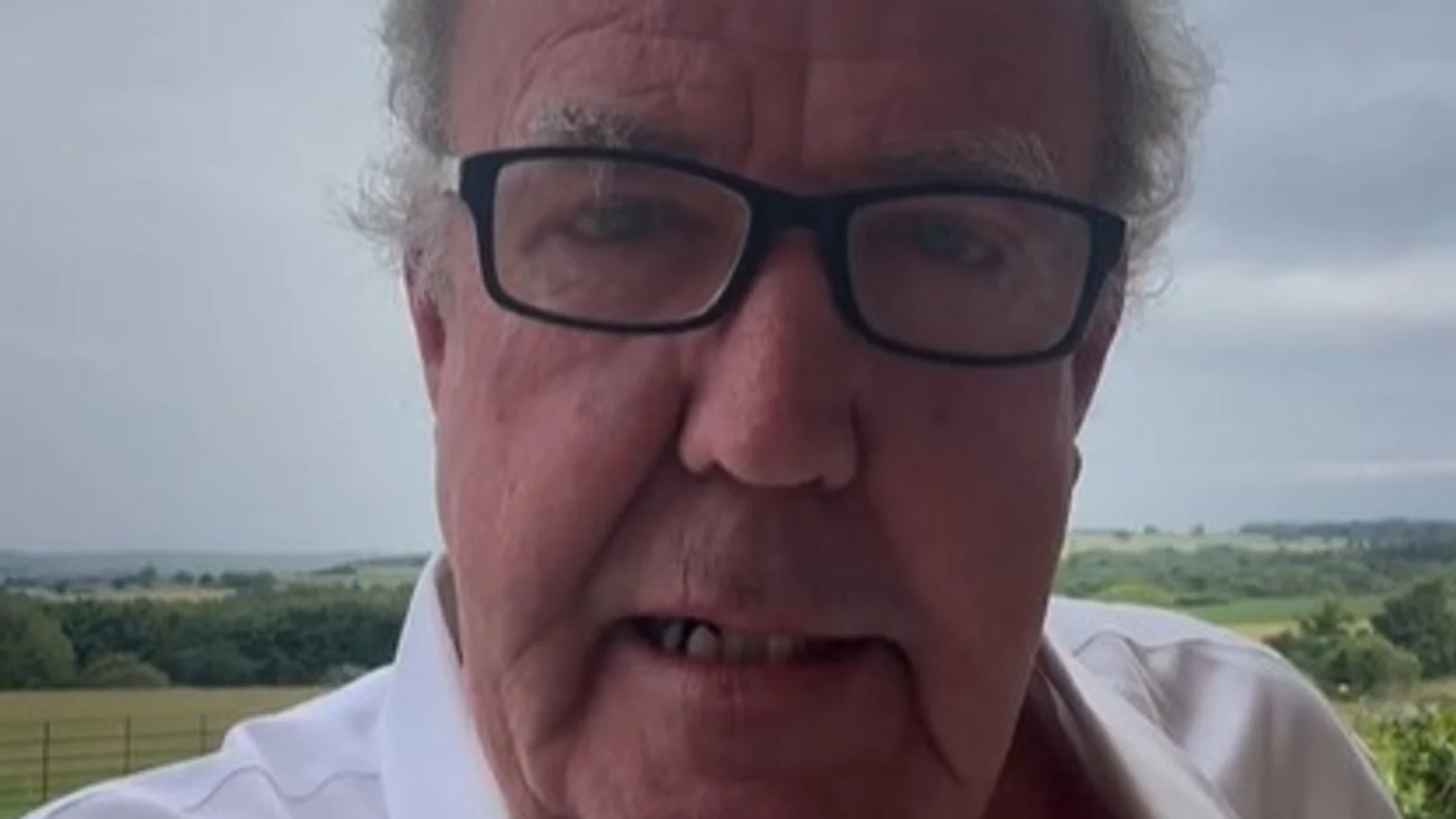 A close-up portrait of Jeremy Clarkson wearing glasses, looking serious, with a blurred green landscape and cloudy sky in the background.