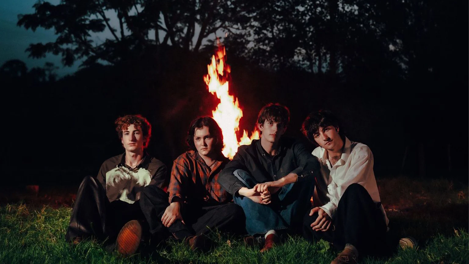 Four young men sit on green grass at night in front of a large, brightly burning bonfire, looking at the camera.