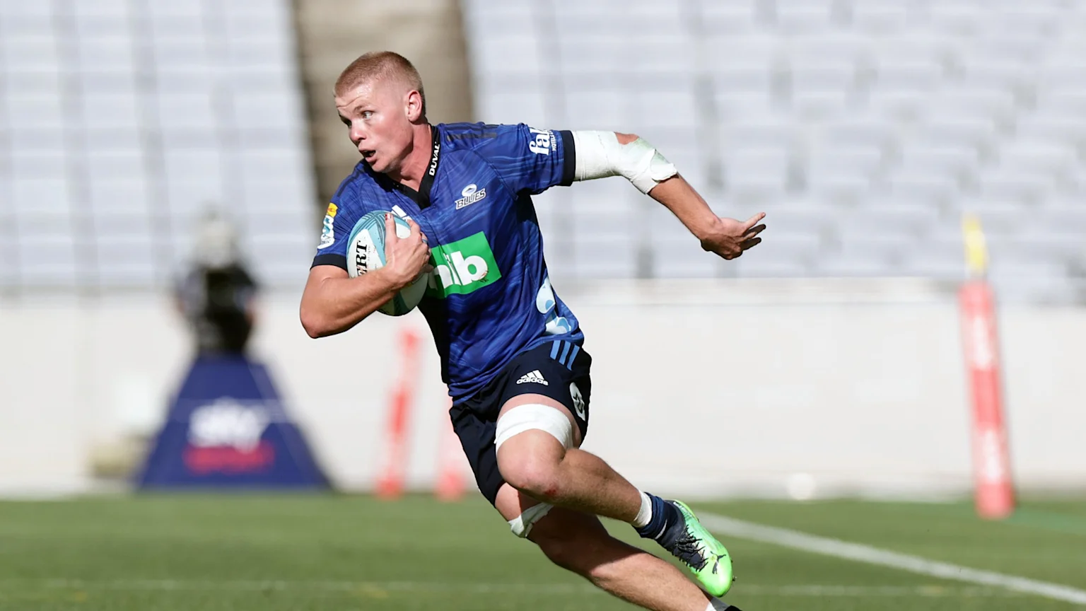A blonde male rugby player in a blue jersey sprints across a green field, clutching a rugby ball, with stadium seats blurred in the background.
