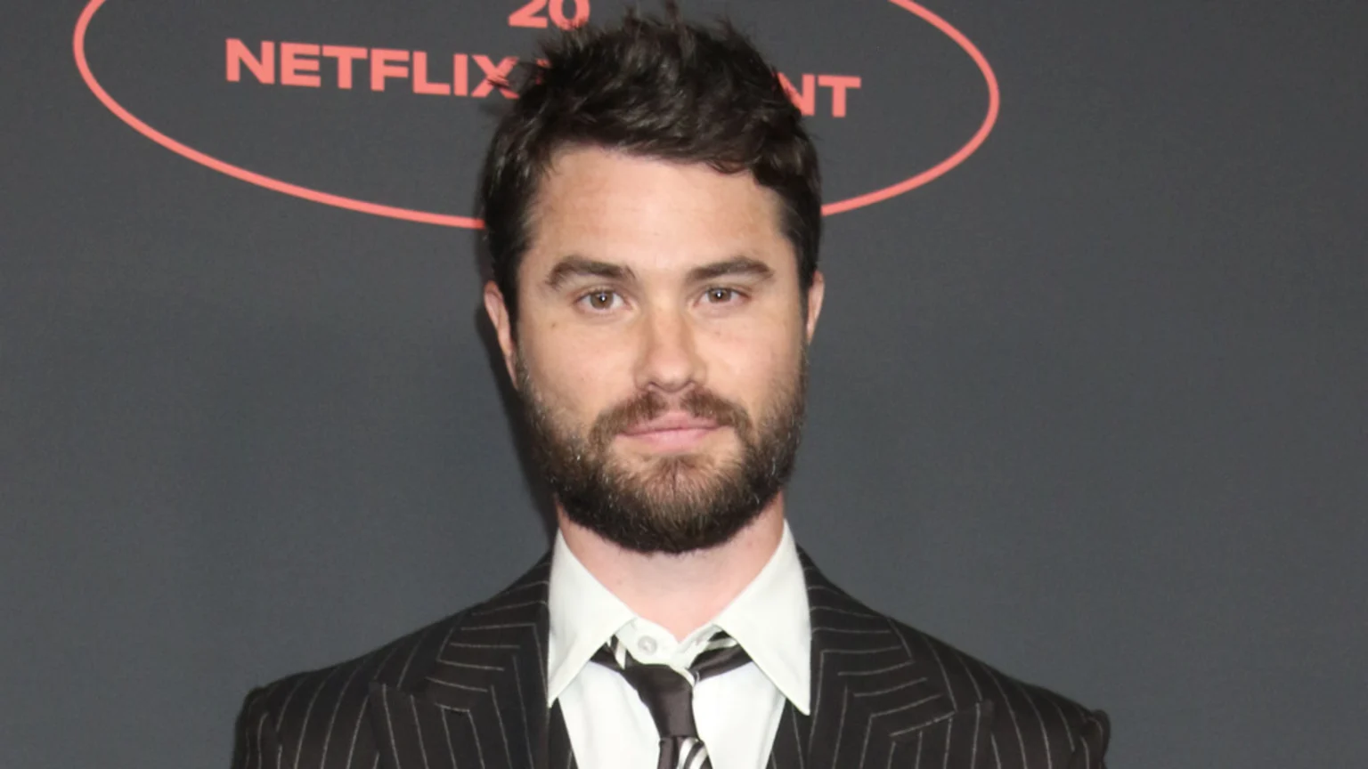 A man with a beard, short dark hair, wearing a pinstripe suit, white shirt, and striped tie, looks directly at the camera with a Netflix logo partly visible in the background.