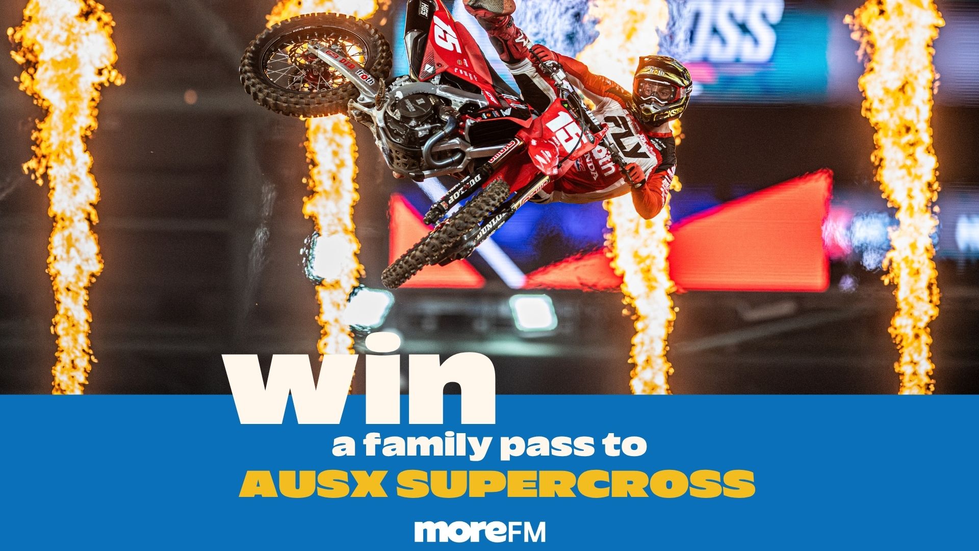 A motocross rider in red and black gear jumps a dirt bike mid-air between two columns of fire, with a blue banner at the bottom announcing "Win a family pass to AUSX SUPERCROSS moreFM."