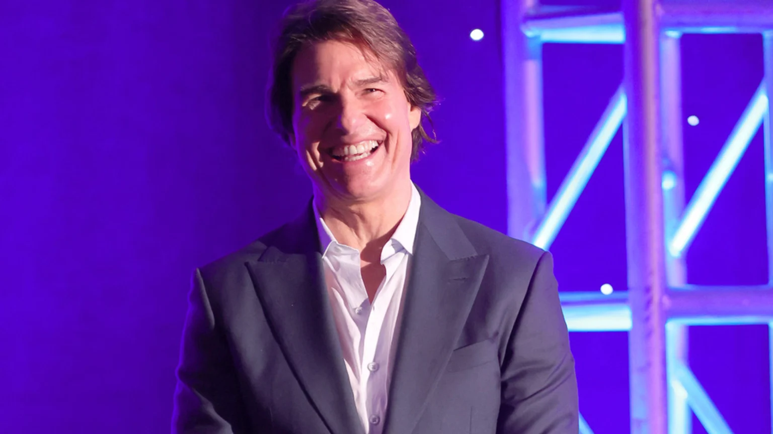 Tom Cruise smiles broadly, wearing a dark suit jacket and a white open-collar shirt, against a vibrant purple background with bright stage lights.